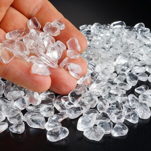 Op de afbeelding: Een hand die heldere kwarts kristalschilfers vasthoudt, met een stapel vergelijkbare kristallen op de achtergrond. De kristallen zijn onregelmatig van vorm en doorschijnend, waardoor ze licht reflecteren. De achtergrond is zwart.