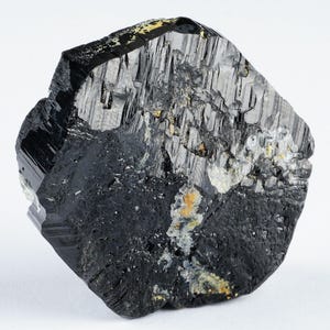 May include: A raw, black tourmaline crystal with a rough, textured surface. The crystal has a dark, glossy appearance with areas of lighter gray and yellow mineral deposits. The stone is set against a plain white background.