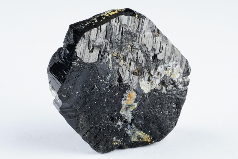May include: A raw, black tourmaline crystal with a rough, textured surface. The crystal has a dark, glossy appearance with areas of lighter gray and yellow mineral deposits. The stone is set against a plain white background.