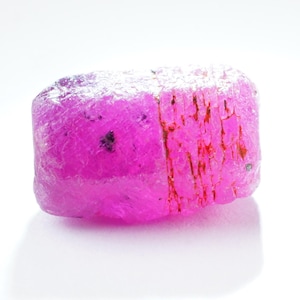 Könnte beinhalten: Ein leuchtend rosa, grob geschliffener Edelstein mit kristalliner Struktur. Der Stein hat eine tiefrosa Farbe mit einem leicht dunkleren, rötlichen Band, das durch ihn verläuft.