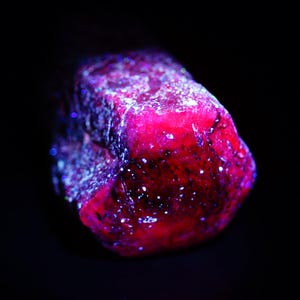 Op de afbeelding: Een close-up van een ruwe, ongeslepen edelsteen, voornamelijk rood met vleugjes paars en wit. De steen heeft een getextureerd oppervlak tegen een zwarte achtergrond, wat een dramatisch contrast creëert. De belichting benadrukt de kristalstructuur.