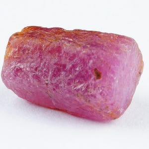 Könnte beinhalten: Ein roher, rosafarbener Rubin-Edelstein mit einem leicht orangefarbenen Farbton. Der Rubin ist ein natürlicher, unpolierter Stein mit einer rauen, unregelmäßigen Form.