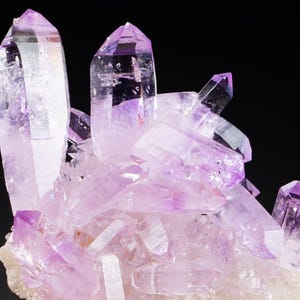 Könnte beinhalten: Eine Ansammlung von durchscheinenden, hellvioletten Amethystkristallen mit scharfen, spitzen Enden. Die Mineralformation weist mehrere vertikale Spitzen unterschiedlicher Höhe auf, die aus einer hellen, strukturierten Basis herauswachsen.