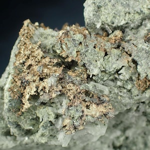 May include: Close-up of a mineral specimen featuring a mix of colors and textures. The piece showcases a matrix of pale green and gray rock with crystalline structures. Metallic, bronze-colored formations are clustered throughout, creating a striking contrast.