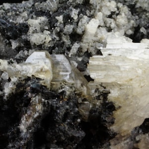May include: A close-up of a rock with white crystals. The crystals are clustered together and have a translucent appearance. The rock is dark grey and has a rough texture.