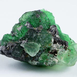May include: A raw, uncut green fluorite mineral specimen. The crystalline structure displays a vibrant emerald green hue with areas of black and silver inclusions. The rough stone is set against a white background.