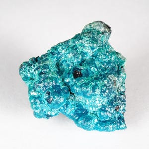 May include: A vivid turquoise mineral specimen with a textured surface. The crystalline structure features small, rounded formations and dark inclusions. The irregular shape highlights its natural geological origins, presenting a captivating display of colour and form.