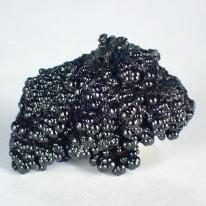 May include: A dark, metallic mineral specimen with a bubbly, rounded surface. The colour is a deep, reflective black, with a slightly uneven texture. The mineral appears to be a cluster of small, spherical formations.