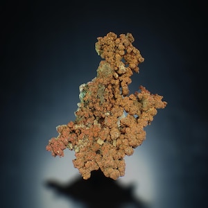 Peut inclure: Gros plan sur un spécimen de minéral de cuivre. L'échantillon présente une surface rugueuse et texturée avec un mélange de teintes brun rougeâtre et vertes. La forme du minéral est irrégulière et agglomérée, sur fond bleu foncé.
