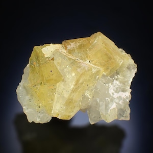 May include: A mineral specimen with a cluster of translucent, cubic crystals. The crystals are a pale yellow with green undertones, set against a dark backdrop. The mineral's structure is crystalline. Measurements are in centimetres.