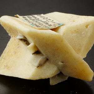 May include: A pale yellow mineral specimen with a complex, geometric structure. The mineral has a rough, textured surface and a small label is attached. The overall shape is angular and multi-faceted. The specimen is a light colour.