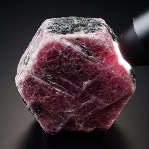 May include: A large, faceted, pink ruby crystal with black inclusions. The crystal is illuminated by a torch.