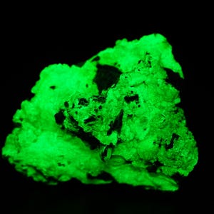 Può includere: Un campione di minerale verde brillante su sfondo nero. Il minerale presenta una superficie ruvida e strutturata con macchie scure e fessure. Il colore verde brillante suggerisce fluorescenza in determinate condizioni di illuminazione.