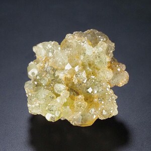 Può includere: Un gruppo di cristalli verde-giallo chiaro con una struttura cristallina scintillante. La formazione minerale ha una superficie ruvida e irregolare ed è posta su uno sfondo scuro. I cristalli hanno una qualità traslucida.
