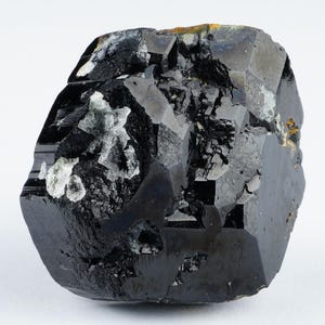 May include: A faceted, black gemstone with a glossy surface. The mineral has a complex, irregular shape with areas of lighter, crystalline inclusions. The overall appearance is dark and reflective, with a few small patches of gold-colored material.