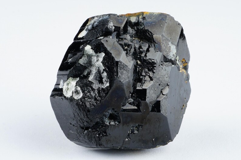 May include: A faceted, black gemstone with a glossy surface. The mineral has a complex, irregular shape with areas of lighter, crystalline inclusions. The overall appearance is dark and reflective, with a few small patches of gold-colored material.