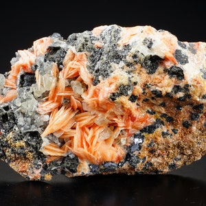 K&ouml;nnte beinhalten: Ein Cluster aus orangefarbenen und wei&szlig;en Kristallen auf einer dunkelgrauen Matrix. Die Kristalle sind scharf und haben eine geschichtete Struktur.