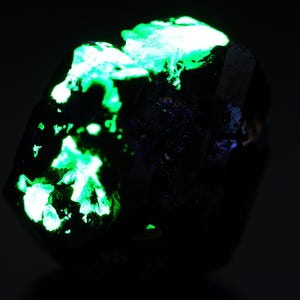 May include: A dark, irregularly shaped mineral specimen with bright, glowing green patches. The contrast between the dark base and the vibrant green highlights the unique texture and crystalline structure of the rock. The image is set against a dark background.