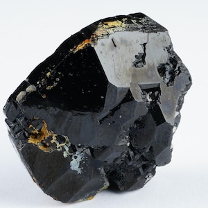 May include: A raw, black tourmaline crystal with a faceted, geometric shape. The surface has a glossy sheen with areas of silver and gold-colored inclusions. The crystal is set against a white background.