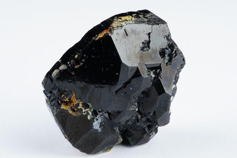 May include: A raw, black tourmaline crystal with a faceted, geometric shape. The surface has a glossy sheen with areas of silver and gold-colored inclusions. The crystal is set against a white background.