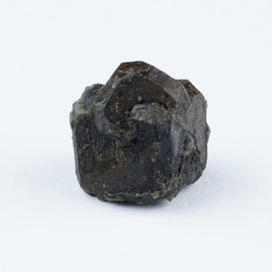 May include: A dark brown, rough-textured mineral specimen against a white background. The stone has a crystalline structure with visible facets and a slightly greenish hue in some areas. The overall shape is irregular and angular.