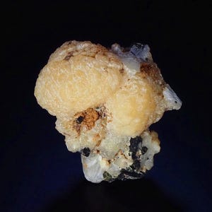 以下が含まれることがあります： 黒い背景に、丸みを帯びた淡い黄色の塊が特徴の鉱物標本。黒と茶色のインクルージョンを持つベースの上にあります。標本は、粗く、質感のある表面を持っています。