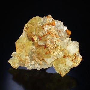 May include: A mineral specimen with a cluster of translucent, cubic crystals in shades of yellow and clear. The crystals have patches of orange and brown, set against a black background. The specimen is a natural formation.