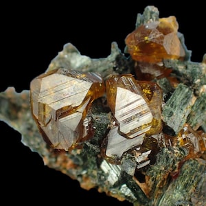 Peut inclure: Gros plan sur un amas de cristaux. Les cristaux sont d'une couleur ambre translucide avec des formes géométriques nettes. Ils sont posés sur un fond de formations minérales vert foncé en forme d'aiguilles et un fond noir. Les cristaux ont un aspect réfléchissant et vitreux.