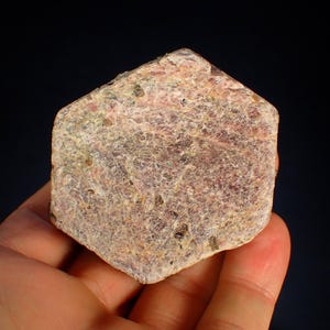 Peut inclure: Une main tenant une pierre précieuse brute, de couleur rouge-rosé. La pierre présente une surface texturée avec des inclusions plus foncées et une forme irrégulière. Le fond est sombre.