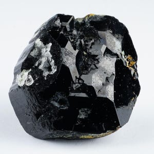 May include: A raw, black tourmaline crystal with a glossy, faceted surface. The gemstone has a rough, irregular shape with areas of lighter, gray-white mineral inclusions. The crystal is set against a plain white background.