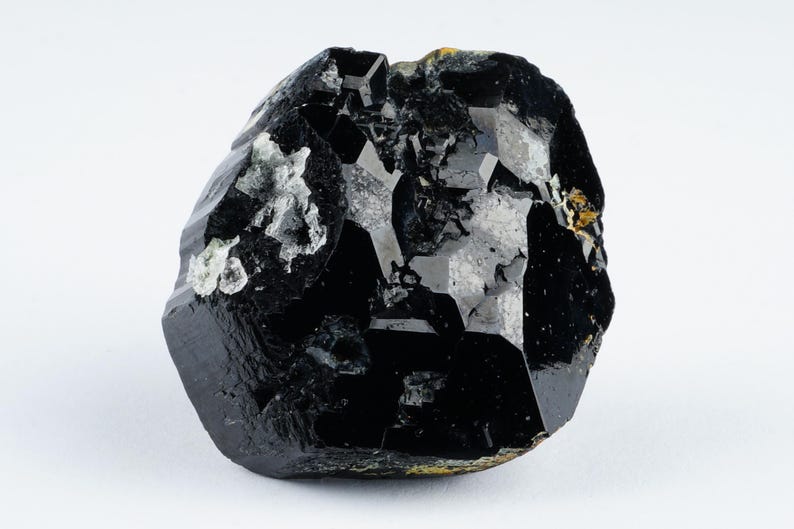 May include: A raw, black tourmaline crystal with a glossy, faceted surface. The gemstone has a rough, irregular shape with areas of lighter, gray-white mineral inclusions. The crystal is set against a plain white background.
