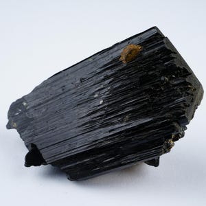 Può includere: Un cristallo nero striato, grezzo e ruvido con una piccola inclusione marrone dorata. Il cristallo è un esemplare naturale grezzo.