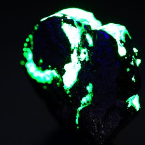 May include: A dark, textured mineral specimen with bright green and white fluorescent patches. The glowing areas contrast against the dark background, highlighting the unique geological features of the rock. The overall effect is striking.