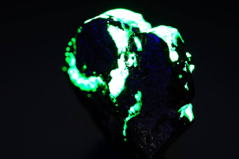 May include: A dark, textured mineral specimen with bright green and white fluorescent patches. The glowing areas contrast against the dark background, highlighting the unique geological features of the rock. The overall effect is striking.