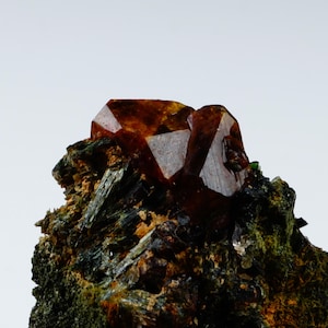 Könnte beinhalten: Ein rohes Mineralexemplar mit einer Ansammlung durchscheinender, tief bernsteinfarbener Kristalle mit scharfen, geometrischen Facetten. Die Kristalle sind in eine dunkle, texturierte Matrix aus faserigen, waldgrünen und anthrazitfarbenen Mineralien eingebettet.