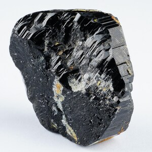 May include: A raw, natural black tourmaline crystal with a rough, textured surface. The gemstone has a dark, glossy appearance with visible striations and some lighter-colored inclusions. The crystal is displayed against a white background.