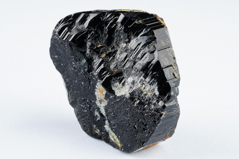 May include: A raw, natural black tourmaline crystal with a rough, textured surface. The gemstone has a dark, glossy appearance with visible striations and some lighter-colored inclusions. The crystal is displayed against a white background.