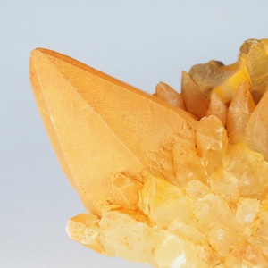 Può includere: Primo piano di un gruppo di cristalli di quarzo dorato. I cristalli hanno una superficie ruvida e strutturata e un colore ambrato caldo. Il punto focale è un grande cristallo appuntito, con cristalli più piccoli che lo circondano. Lo sfondo è un azzurro tenue.