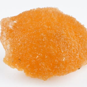 May include: A close-up of an orange mineral specimen. The surface is rough and textured, with small, granular structures. The shape is irregular, with a slightly rounded top and a jagged edge. The colour is a vibrant orange.