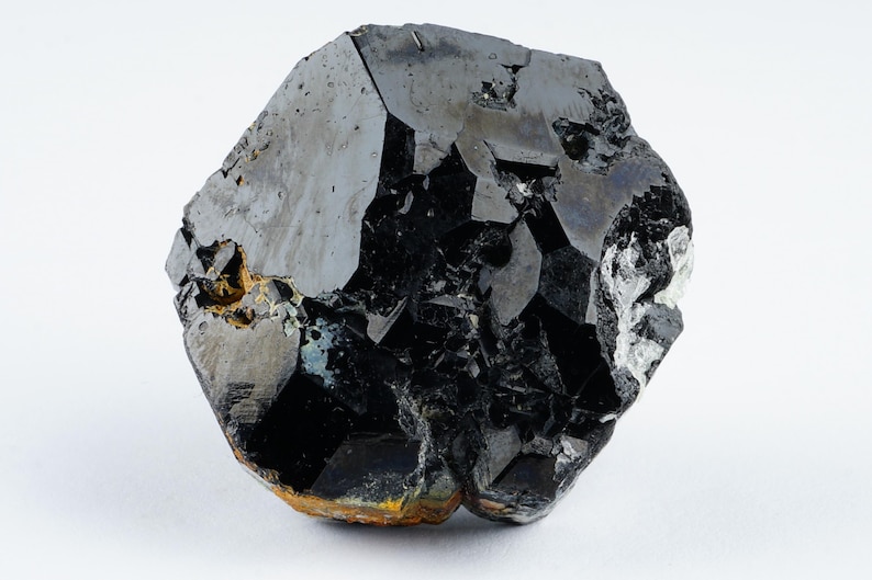 May include: A close-up of a large, irregularly shaped black mineral specimen. The surface has a glossy sheen with areas of lighter gray and hints of gold and white. The mineral is set against a plain white background.