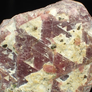 May include: A close-up image of a natural mineral specimen. The stone displays a blend of colours, including pale pink, cream, and dark reddish-brown. Geometric patterns are visible across the surface, with small dark inclusions scattered throughout. The texture appears rough and crystalline.