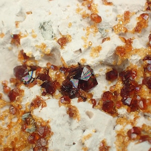 Puede incluir: Primer plano de un espécimen mineral con una matriz de roca blanca y grupos de pequeños cristales de color marrón rojizo brillante. También se ven varios cristales más grandes, facetados y de color rojo oscuro.