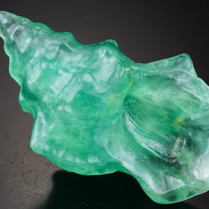 Op de afbeelding: Een doorschijnende, zeegroene fluorietkristalformatie. De gepolijste steen heeft een wervelende, organische vorm, met een geribbelde, puntige punt en een glad, vloeiend lichaam. Het kristal wordt tegen een donkere achtergrond gepresenteerd.