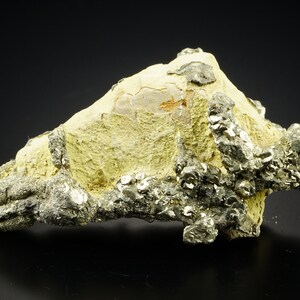 Può includere: Un gruppo di cristalli di pirite grigio metallico su una matrice rocciosa gialla. I cristalli sono nitidi e ben definiti, con un lucentezza metallica. La roccia è ruvida e testurizzata.