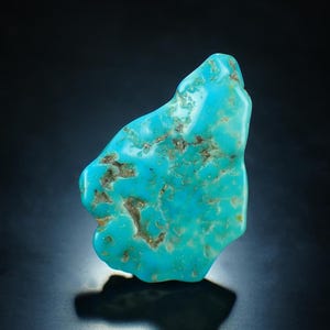 Può includere: Una vibrante pietra preziosa di turchese, di forma irregolare e con superficie lucida. La pietra mostra una sorprendente miscela di tonalità blu e verdi brillanti, accentuata da motivi a matrice marrone. La gemma è posta su uno sfondo scuro.