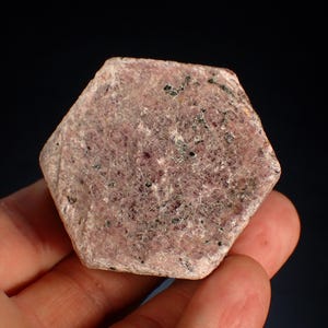May include: A hexagonal, rough-cut ruby gemstone with a pinkish-red hue. The surface has a textured appearance with small dark inclusions. The gemstone is held in a hand against a dark background. This natural gemstone is a unique piece.