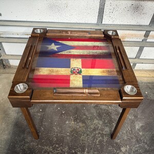 May include: A wooden domino table featuring the flags of Puerto Rico and the Dominican Republic. The table has a dark brown finish, four stainless steel cup holders, and slots for dominoes. The table is supported by four wooden legs.
