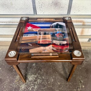May include: A square wooden table with a glass top featuring a graphic of a Cuban flag, a bottle of Havana Club rum, and a glass of amber liquid. The table includes four metal cup holders and integrated slots for cigars on each side.