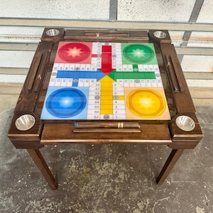 May include: A wooden Ludo board game table with a dark brown finish. The game board features red, green, blue, and yellow sections. It has built-in cup holders and storage for game pieces. The table is supported by four angled legs.