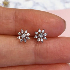 May include: A pair of small, silver-toned stud earrings in a floral design. Each earring features a cluster of clear, sparkling gemstones arranged in a flower shape. The earrings are displayed against a soft pink background.